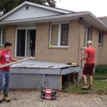 two young men dismantle a grey deck from the back of our tan brick house