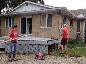 two young men dismantle a grey deck from the back of our tan brick house