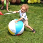 Two year old boy with wild, curly blond hair attempts to hit a beach ball with a kiddie golf club