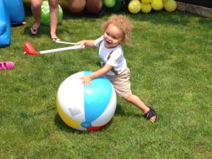 Two year old boy with wild, curly blond hair attempts to hit a beach ball with a kiddie golf club