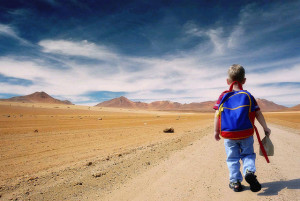 A little boy wearing a blue backpack and carrying a paper bag lunch walks on a dirt road and all you can see for miles ahead is barren desert