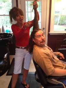 A woman wearing a red top and white shorts holds Derek's ponytail up straight and aims her scissors at it while he looks cartoonishly frightened