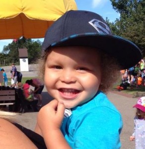 LIttle boy in a light blue shirt and dark blue baseball cap smiling brightly 