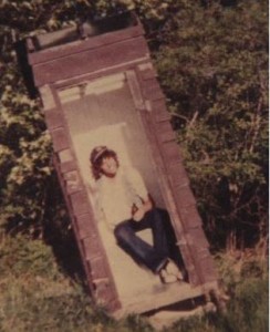 An outhouse leaning to the left, with a lanky teen sitting inside, holding a beer bottle and smiling broadly