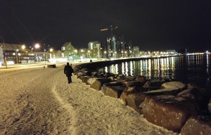 A wide view of the harbour lights on the water and me, mid-frame, walking along the showy shore