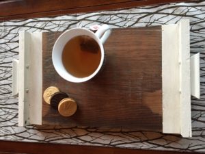 Barnboard tray has unfinished centre with cream-coloured painted handles on either end. Holding a up of tea and a few cookies.