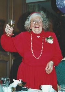 Auntie Barb, wearing a bright red dress, her long grey hair held back by a headband, toasting the camera with a glass of wine