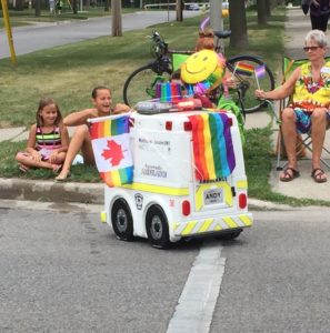 mini ambulance decked out in rainbow colours travels close to a sidewalk where parade watchers sit. 