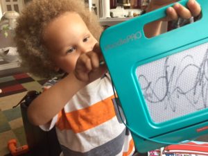 Ryker, with his curly blond hair, holds up the turquoise plastic drawing board with squiggles on it