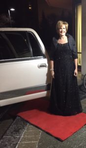 Erin wears a black, sparkly gown as she poses on the red carpet beside a white, stretch limo
