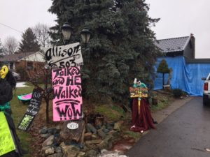 pink, lime green and white lawn signs, figures dressed as zombies, collected on a front lawn with blue tarps flapping on the house in the background