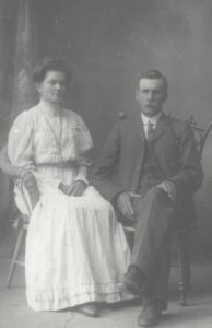 Man and woman, formally dressed, sit side by side on wooden chairs, not smiling
