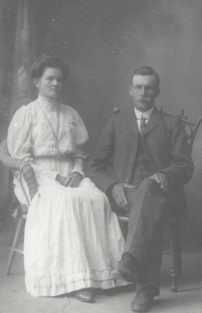Man and woman, formally dressed, sit side by side on wooden chairs, not smiling