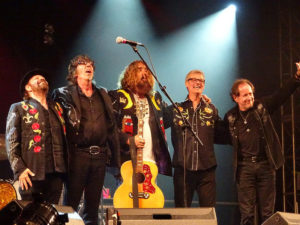 Five musicians arm in arm at the end of a 2016 concert. Tom Wilson is in the middle with a yellow guitar.