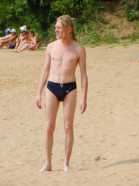 skinny white man with a ponytail and navy bathing suit standing on the sand, looking into the distance