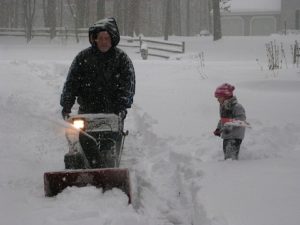 Man snowblowing a driveway in a heavy snowfall while a young boy shovels show nearby
