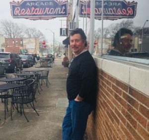 Derek leans against the brick wall of the Arcade restaurant as its neon sign reflects in the window above him