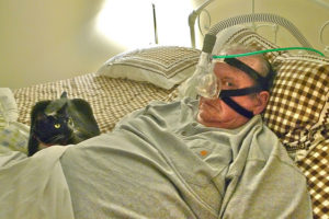 An older man lies in bed wearing a CPAP machine, looking frightened at the camera while his grey cat lays at his side looking at him warily