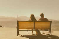 At sunset, a couple sitting on a bench with their dog at their feet, watching the water. They're seen from behind.