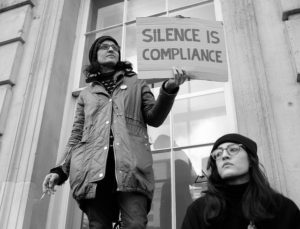 two women outside the US consulate in London England. One holds a sign that reads Silence is Compliance