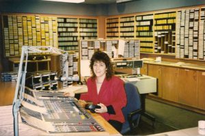 me sitting at the massive control board in the MIX 999 studio at 2 St. Clair West in Toronto. Behind me are racks and racks of carts (cartridges) and CDs. I have permed hair!