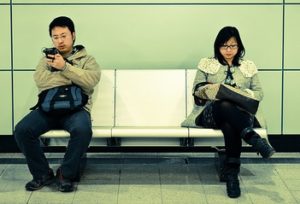 a man and a woman sit at opposite sides of a bench in a Hong Kong subway station. He's looking at his phone and she's reading a book.