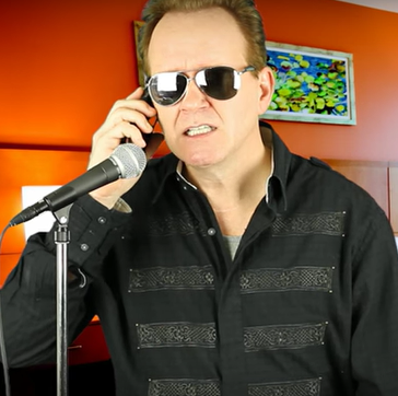 Ed Kelly standing in front of a microphone, wearing mirrored sunglasses, talking on the phone as he does one of his impressions
