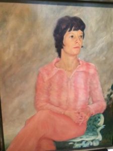 painting of my Mom. She has short, dark hair and is wearing a pink pantsuit, sitting on a white chair with her legs crossed and arms folded, looking off to the left