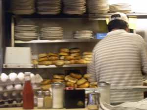 The back of a cook with stacks of plates, eggs and bagels in front of him as he works behind a counter in a diner