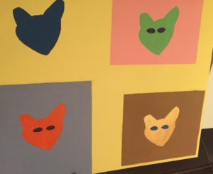 Four-panel painting with a cat head in each. A wide variety of colours used for cats and backgrounds
