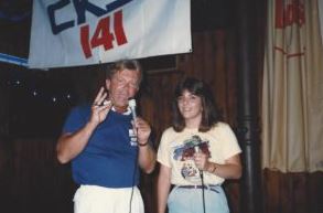 Rich, wearing a blue shirt and white pants, and me, about 22 years old in a yellow shirt with purple grapes on it, below a CKSL banner, holding microphones