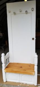 Old door painted white with five crystal doorknobs as coat hangers. A bench at the bottom features railings with newel posts on either side made from an old footboard. The bench top is varnished wood.