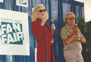 Very thin and frail-looking Tammy Wynette in a long, red blazer and sunglasses speaks on the mic next to George who looks like a typical old man who has money