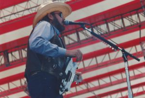 Hank Williams Jr singing on stage in front of a backdrop of an American flag