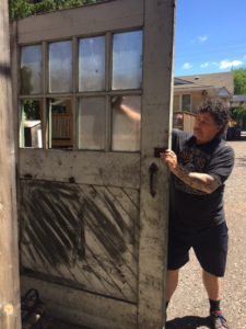 Derek in work clothing holding up a door that's at least eight feet high. It was once white but has black smears of paint on the bottom. The top half is composed of small window panes with two missing. 