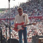 A shot of Parnell playing his guitar from the perspective of backstage. A massive audience can be seen behind him.