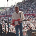 A shot of Parnell playing his guitar from the perspective of backstage. A massive audience can be seen behind him.