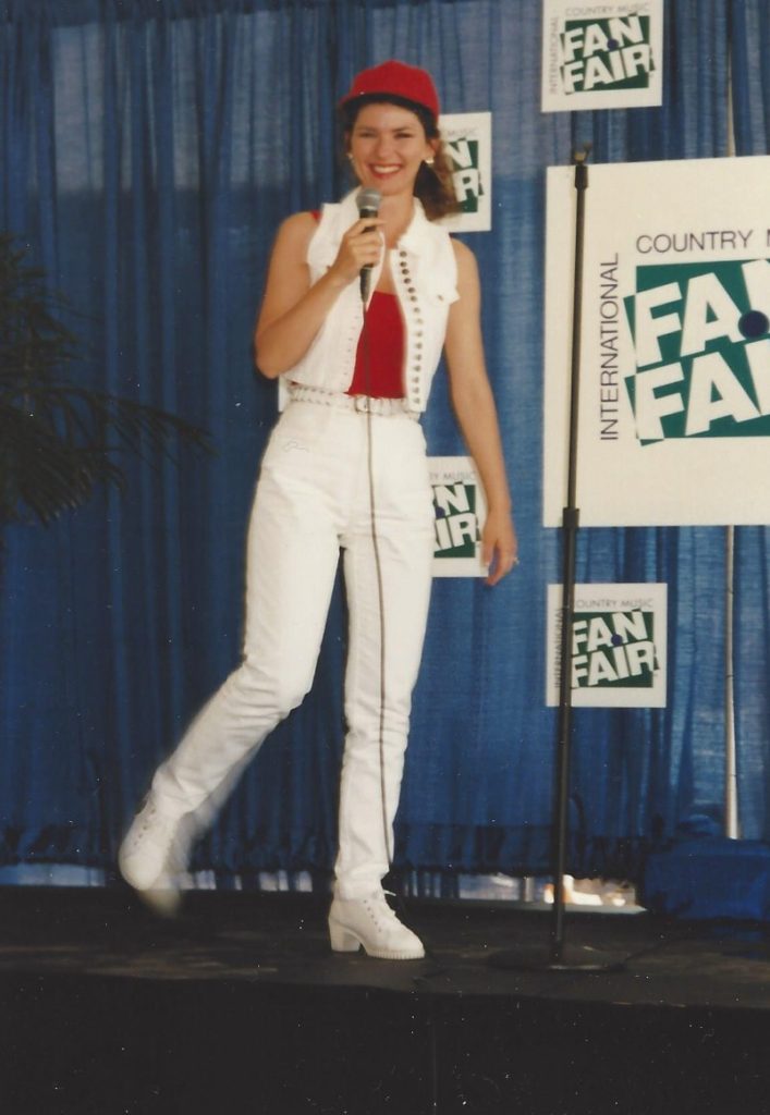 Shania \Twain in white jeans, white sleeveless denim jacket, red top and red hat, looks happy as she walks onto the Fan Fair media stage