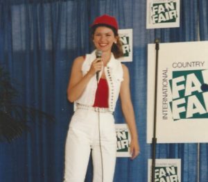 Shania \Twain in white jeans, white sleeveless denim jacket, red top and red hat, looks happy as she walks onto the Fan Fair media stage