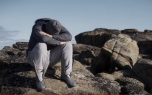 One of the opinionated white men sitting on rocks with his head buried in his lap, deeply unhappy