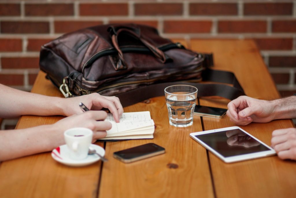 The hands of two people in a cafe, one is taking notes in a note pad and the other is working on an iPad with coffee cups and messenger bags in the background