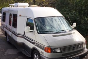 Rialta motorhome looks like a large van. White with tan and brown stripes. The side has a bit of a bump-out and you can see a short air conditioning unit on the top. 