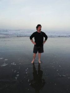 Derek standing at the water's edge on a beautiful, sandy beach. He's wearing shorts and a long-sleeved shirt and the sun is rising behind him