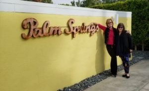 Erin and I arm in arm in front of a yellow wall with metal lettering spelling Palm Springs