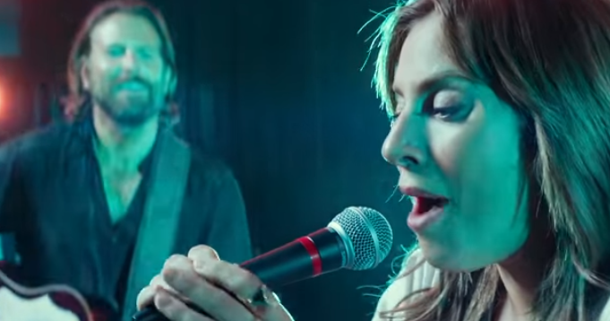 screen shot from A Star is Born. Lady Gaga as Ally performs on stage for the first time while Jack (Bradley cooper) plays guitar and watches in admiration