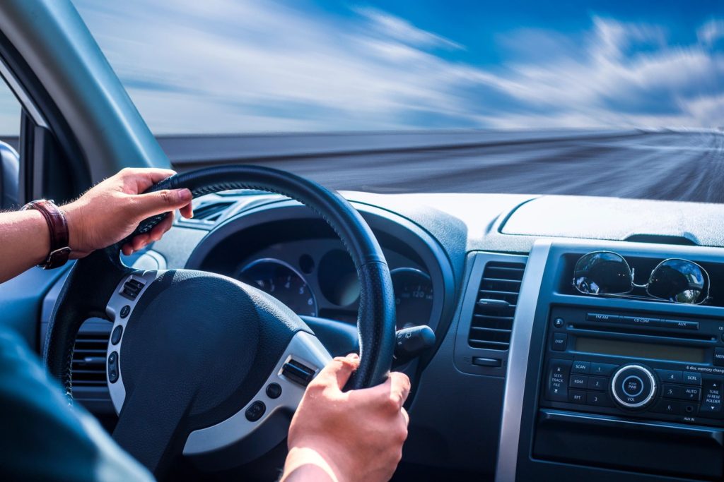 hands on the wheel of a car, and the dashboard, with blue sky and a clear road ahead