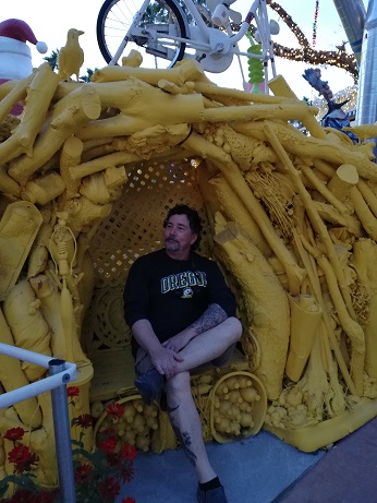 Derek sitting inside a little cave with a bench all made from tree branches and scraps of wood and painted bright yellow