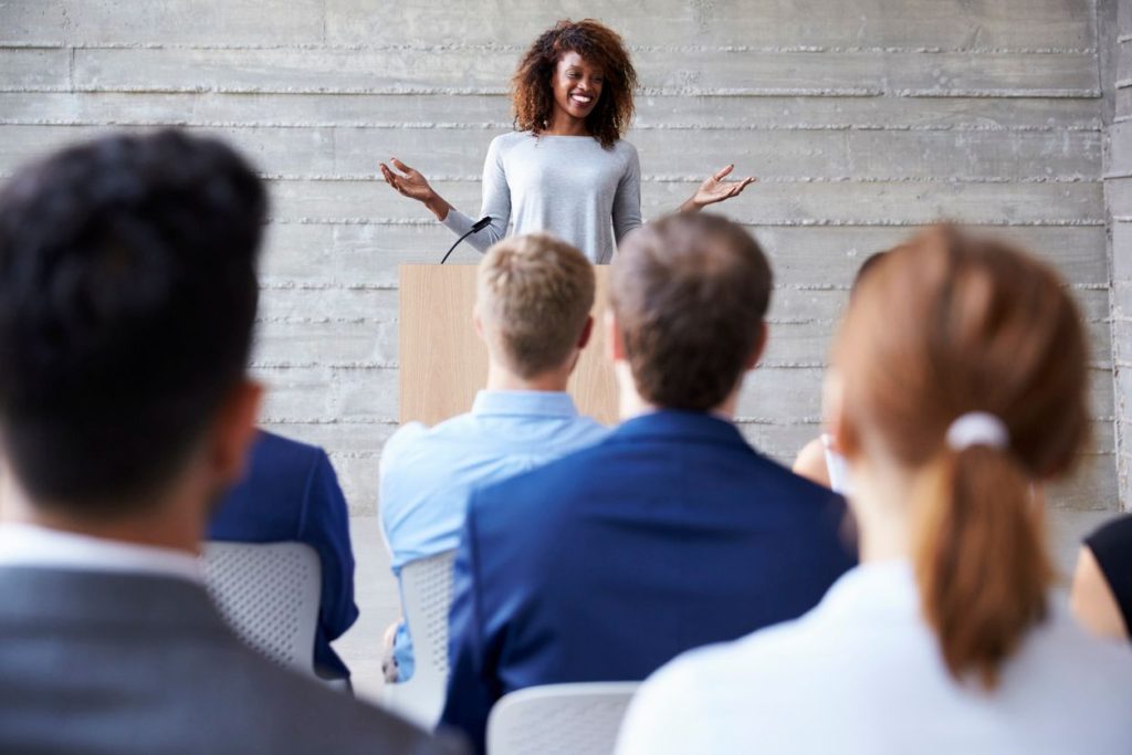 a woman gesturing in front of rows of people as she gives a speech