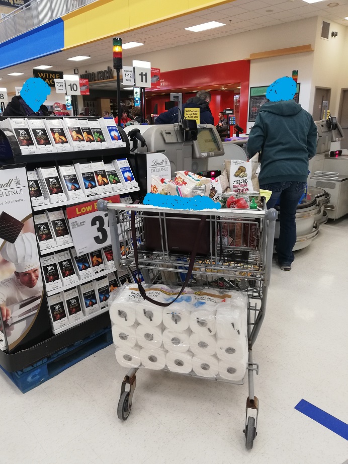 Woman fussing with bags at the self checkout while her purse sits open and unattended a good eight feet behind her, in the shopping cart. She has a massive package of toilet-paper rolls on the rack of the bottom of the cart.