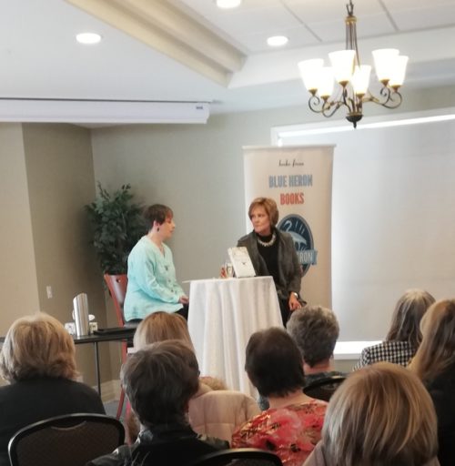 Forty-something woman with very short hair, wearing a long-sleeved mint-green top sitting at a round table covered in a long, white tablecloth, while Erin Davis looks at her, listening to her question. The heads of many attendees are in the bottom of the frame.